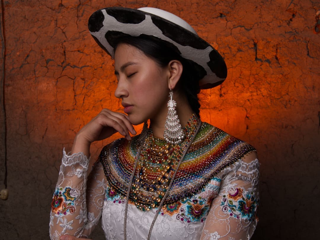 A young Saraguro girl dressed in her traditional attire. She wears a white wool hat with black spots reminiscent of cowhide, silver earrings that drip to her shoulders and a thick necklace of rainbow-colored beads. Her white blouse is adorned with lace and embroidery in primary colors. The background is dimly lit, with soft light highlighting her face and upper body, calling attention to the intricate details of her clothing and jewelry.   