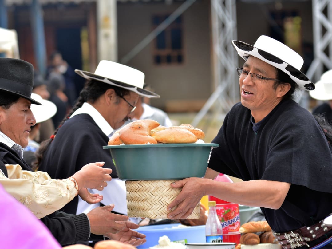 Una persona Saraguro con ropa típica pasa una bandeja llena de pan por una mesa donde otros Saraguros preparan comida.