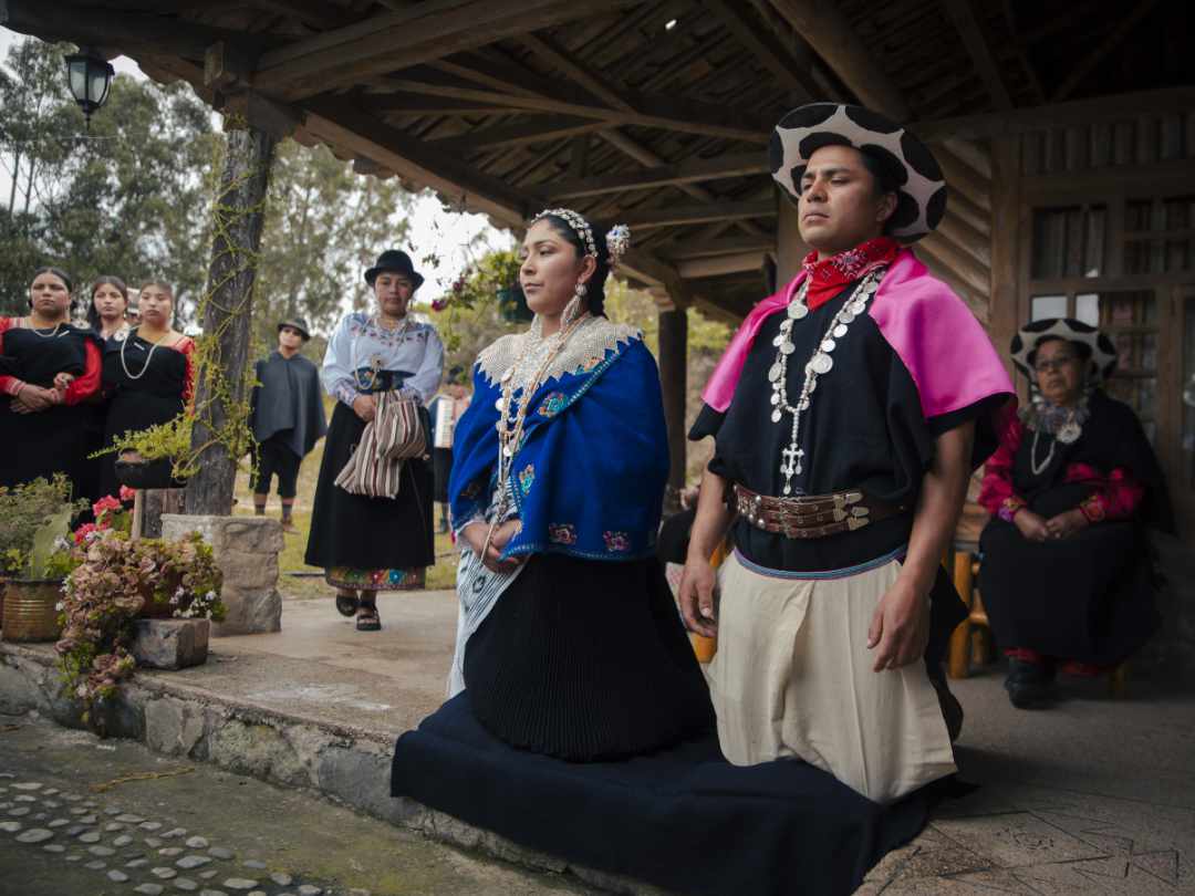 A Saraguro couple kneels in the foreground during a ceremony while their community observes from behind.
