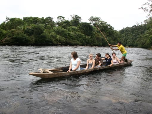 Canoa Tradicional Cinco mujeres jóvenes están sentadas en una canoa tradicional de madera, mientras un miembro de la comunidad de Machacuyacu, ubicado en la posición trasera de la canoa, la guía con un remo, reflejando un momento de convivencia cultural y armonía con la naturaleza.