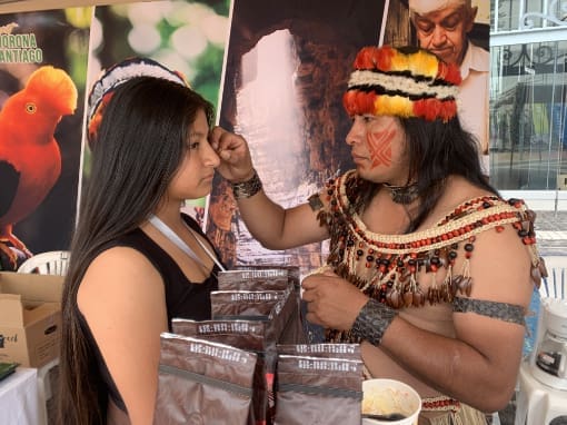 Conexión Shuar Un miembro de la nacionalidad Shuar, vestido con atuendo tradicional que incluye una banda de plumas y un collar, pinta el rostro de un visitante con achiote, en un acto de intercambio cultural y conexión ancestral.