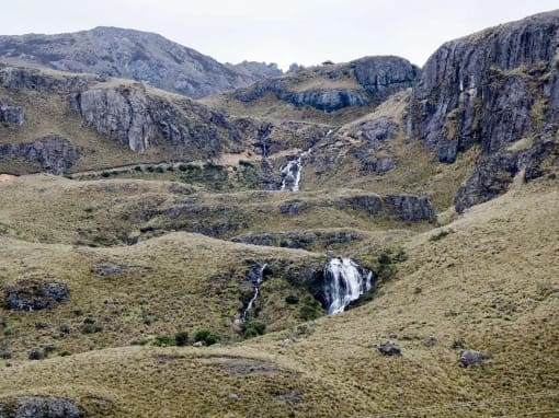 Parque Nacional Las Cajas Cascada en el Parque Nacional Cajas rodeada de vegetación de páramo color marrón dorado, entre grietas rocosas y montañas imponentes.