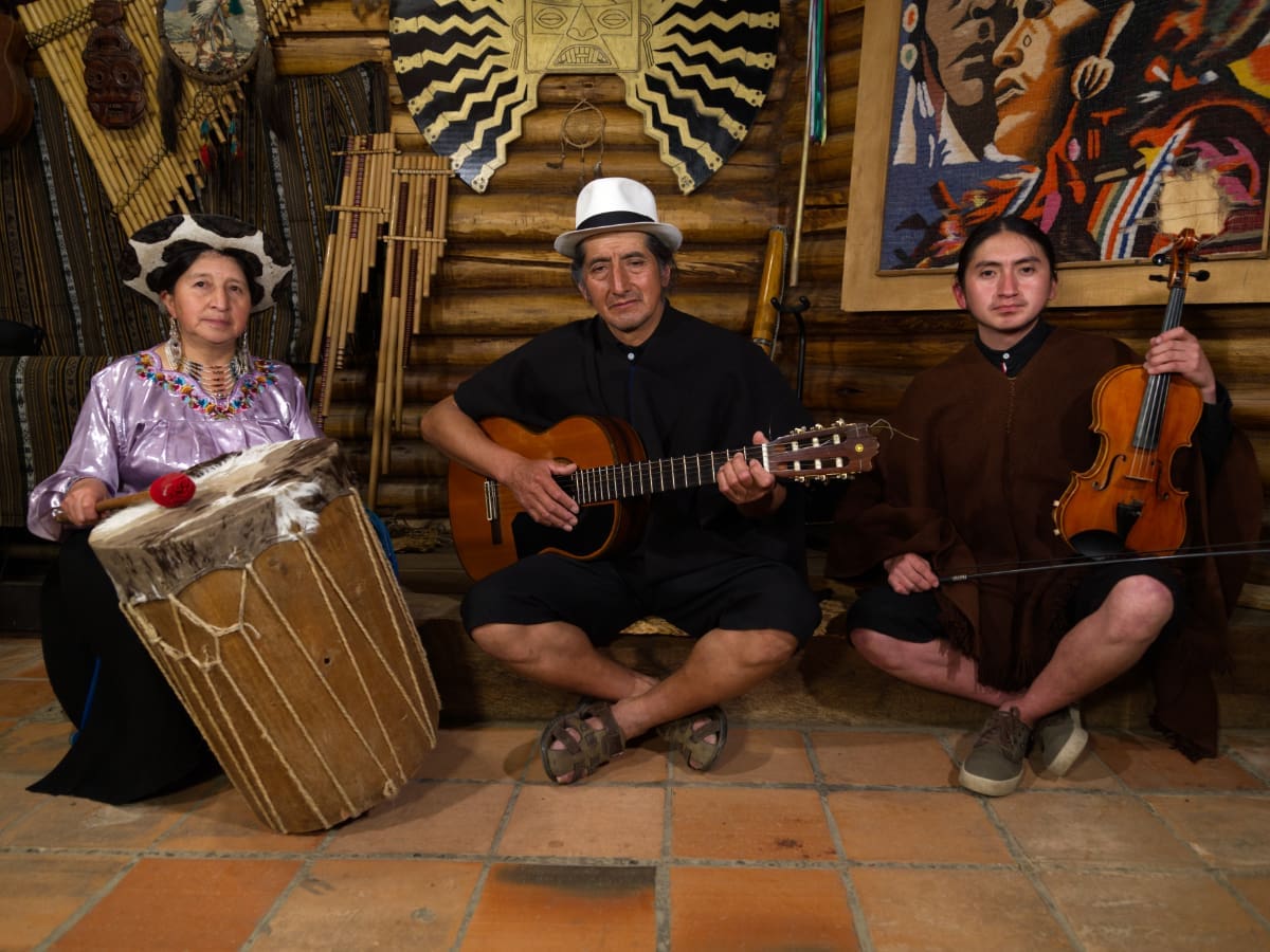 Three musicians with traditional instruments, dressed in the typical Saraguro dress, sitting on the floor.