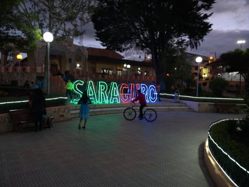 A colorful neon sign with the word 'Saraguro' glows brightly in the park in front of the church at dusk, while children on bicycles appear as silhouettes in profile.
