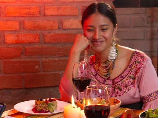 El restaurante Tupay Una mujer vestida con una blusa bordada y aretes tradicionales de Saraguro sonríe mientras le sirven un plato de comida inspirado en la cultura andina durante una cena a la luz de las velas en el restaurante Tupay.