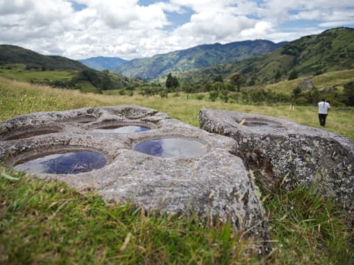 Ancient rocks with carved basins, possibly used for grinding, lie in the middle of a pasture surrounded by mountains, as a man walks in the distance.